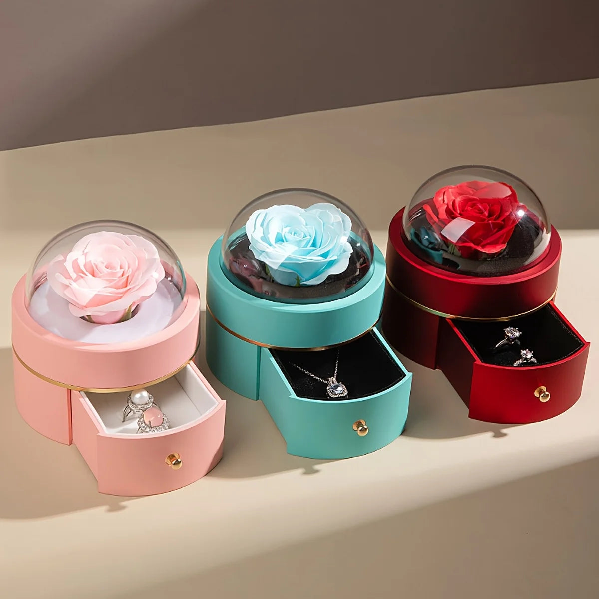 Pink, blue and red round rose jewellery boxes with drawers open showing a ring, a necklace and earrings, roses under clear domes