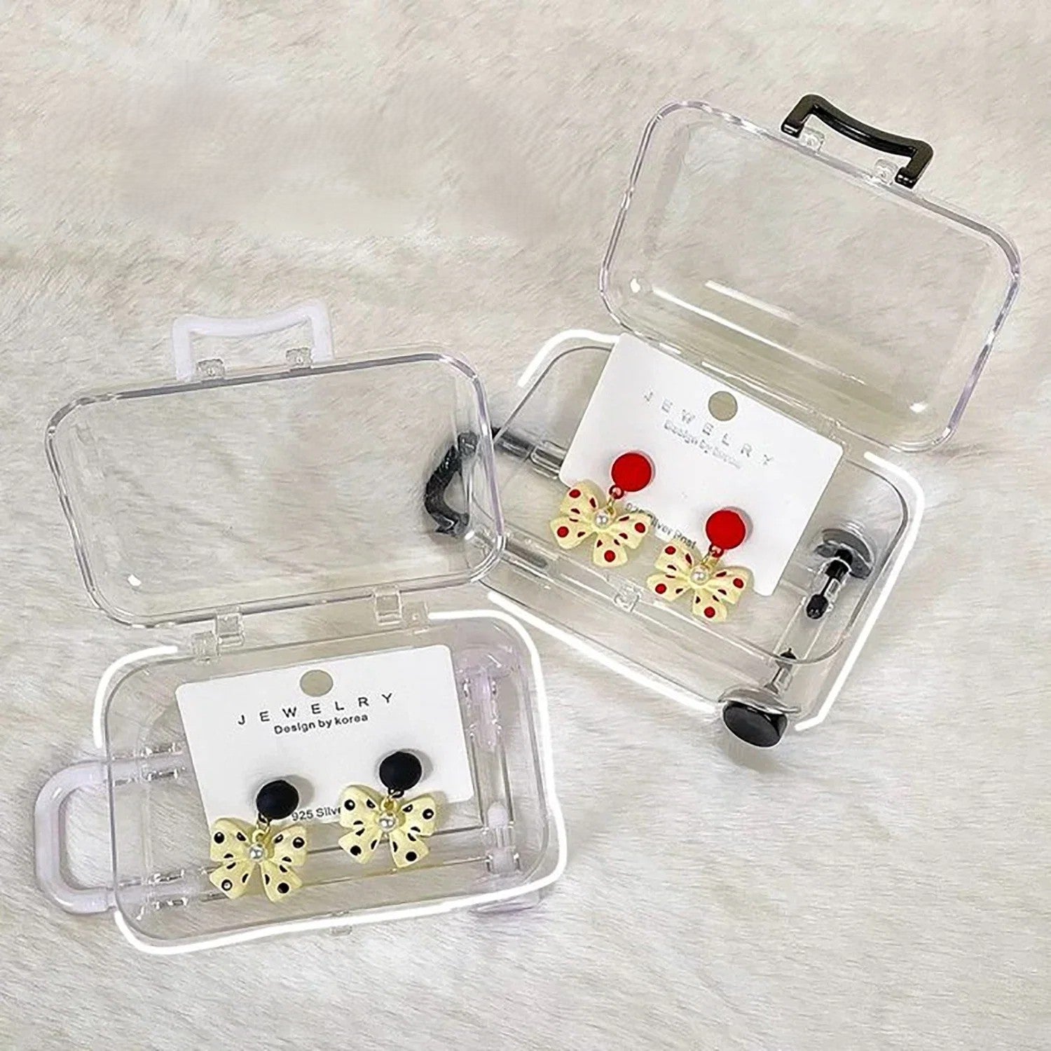 Two transparent acrylic mini suitcase display boxes with one black and one white colour-matched handle, wheels, and latch, displaying earrings.